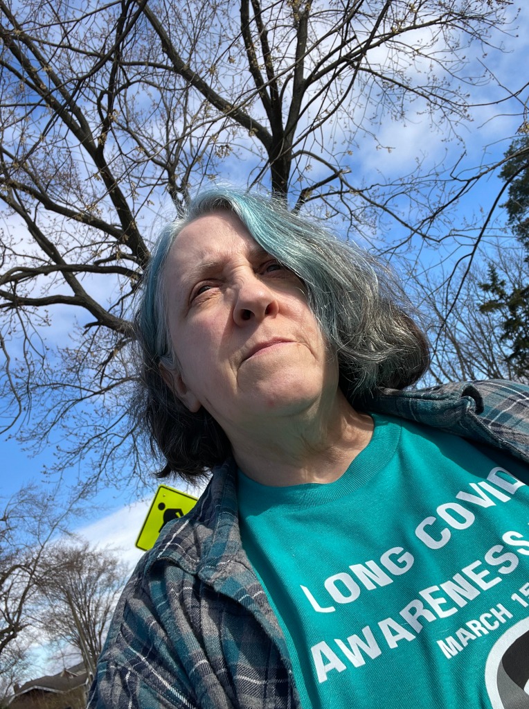Middle-aged white woman with blue hair wearing a teal-blue tshirt which has the text "Long COVID Awareness, March 15." She is outdoors with a blue sky behind her, some bare tree branches and a street-crossing sign. 